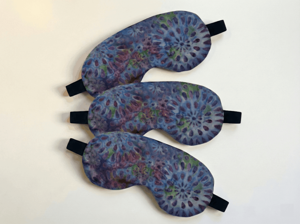 a lavender eye mask three pack. each eye mask has a purple floral tie-die pattern on it, is hand sewn, and has an elastic strap. the eye mask is filled with lavender buds has a subtle natural fragrance.